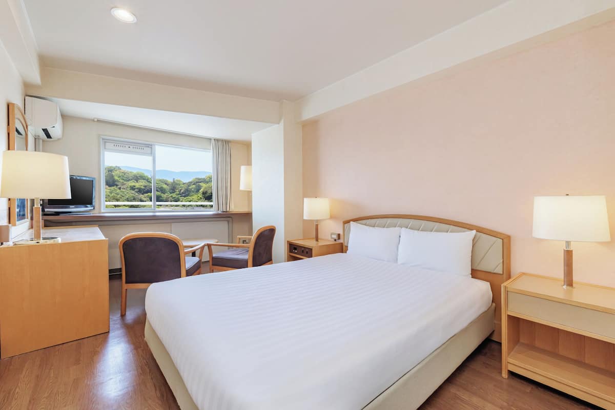 A hotel room with a large bed, a desk with a chair, a TV, and a window with a view of greenery.