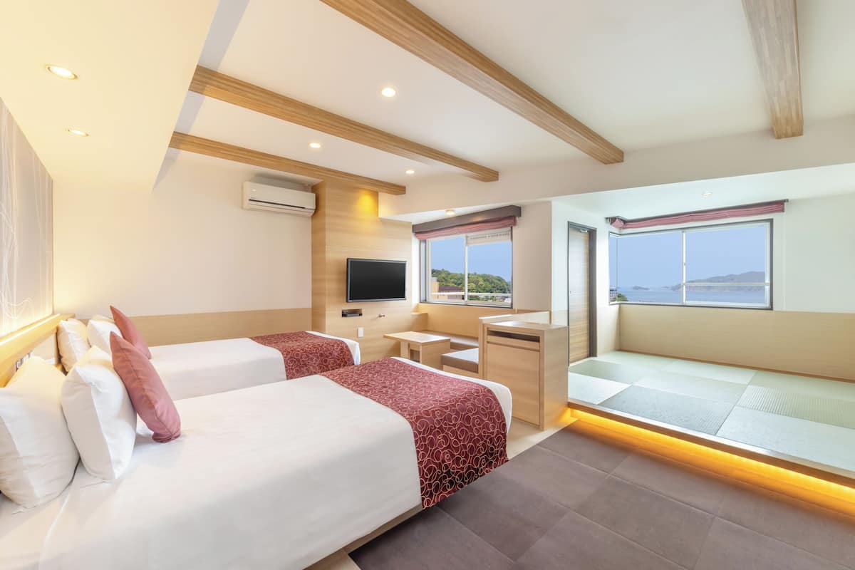 A hotel room with two beds, a desk, a television, and a view of the sea.