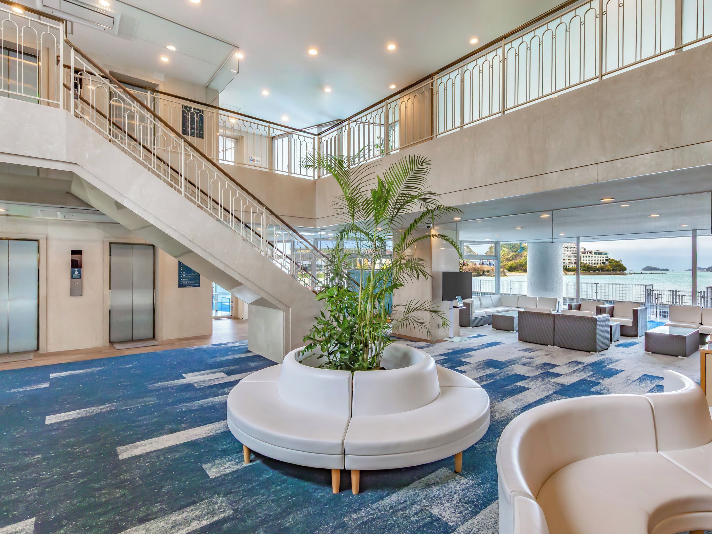 A spacious lobby with a central seating area, a staircase, and a view of the sea.