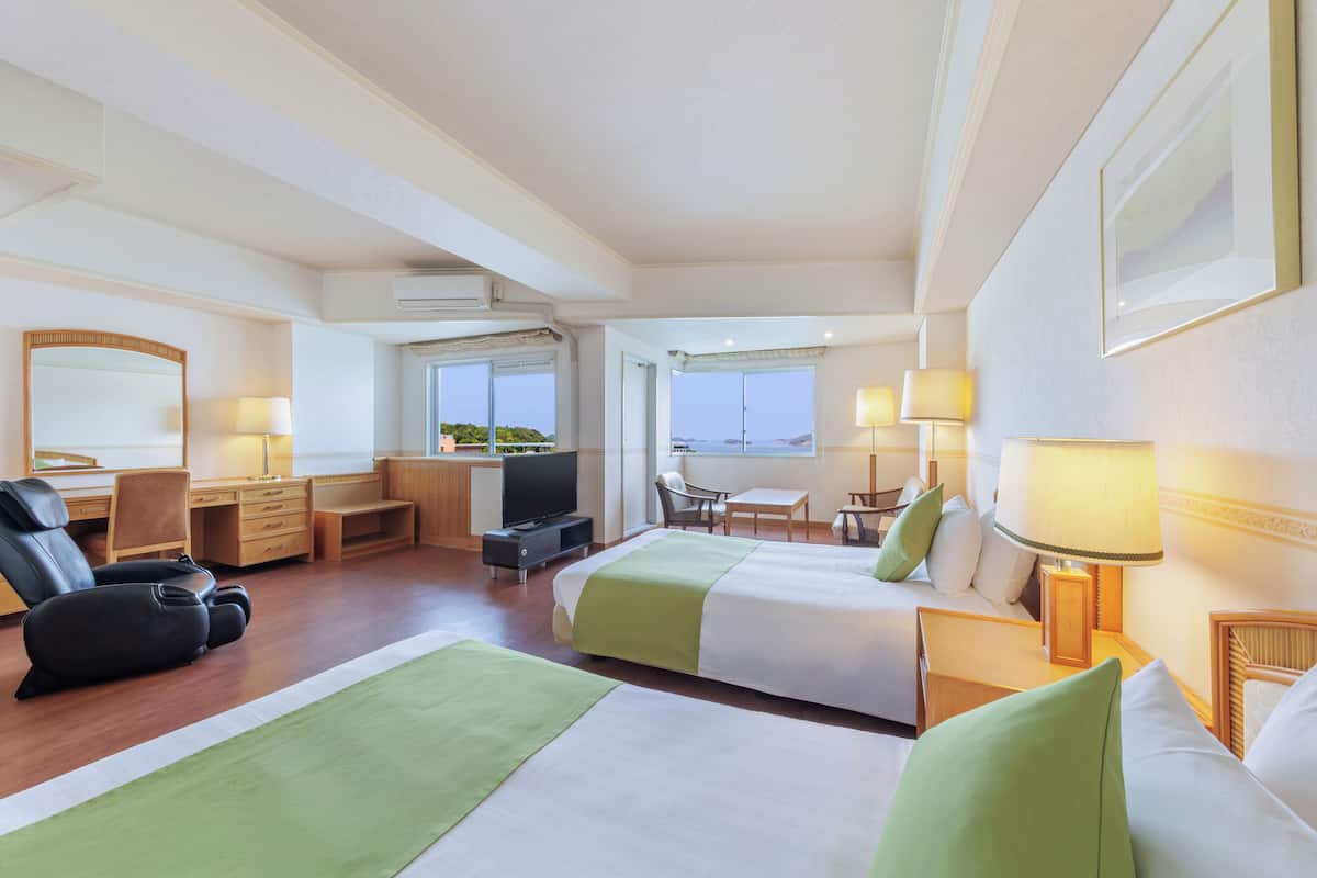 A hotel room with two beds, a desk, a chair, a TV, and a view of the sea.