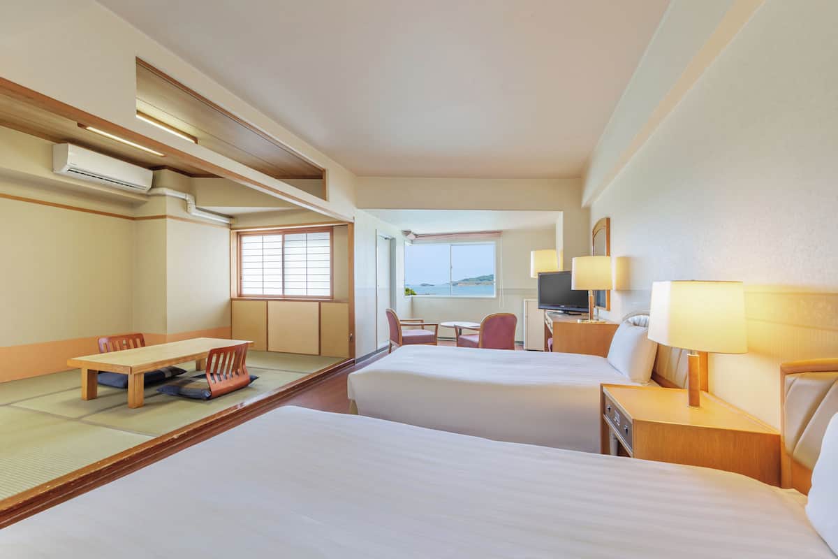 A hotel room with two beds, a dining table, chairs, a TV, and a view of the sea.
