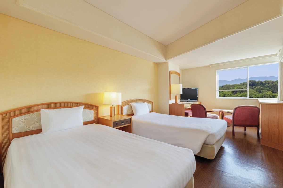 A hotel room with two beds, a desk, a chair, a television, and a window with a view of the mountains.