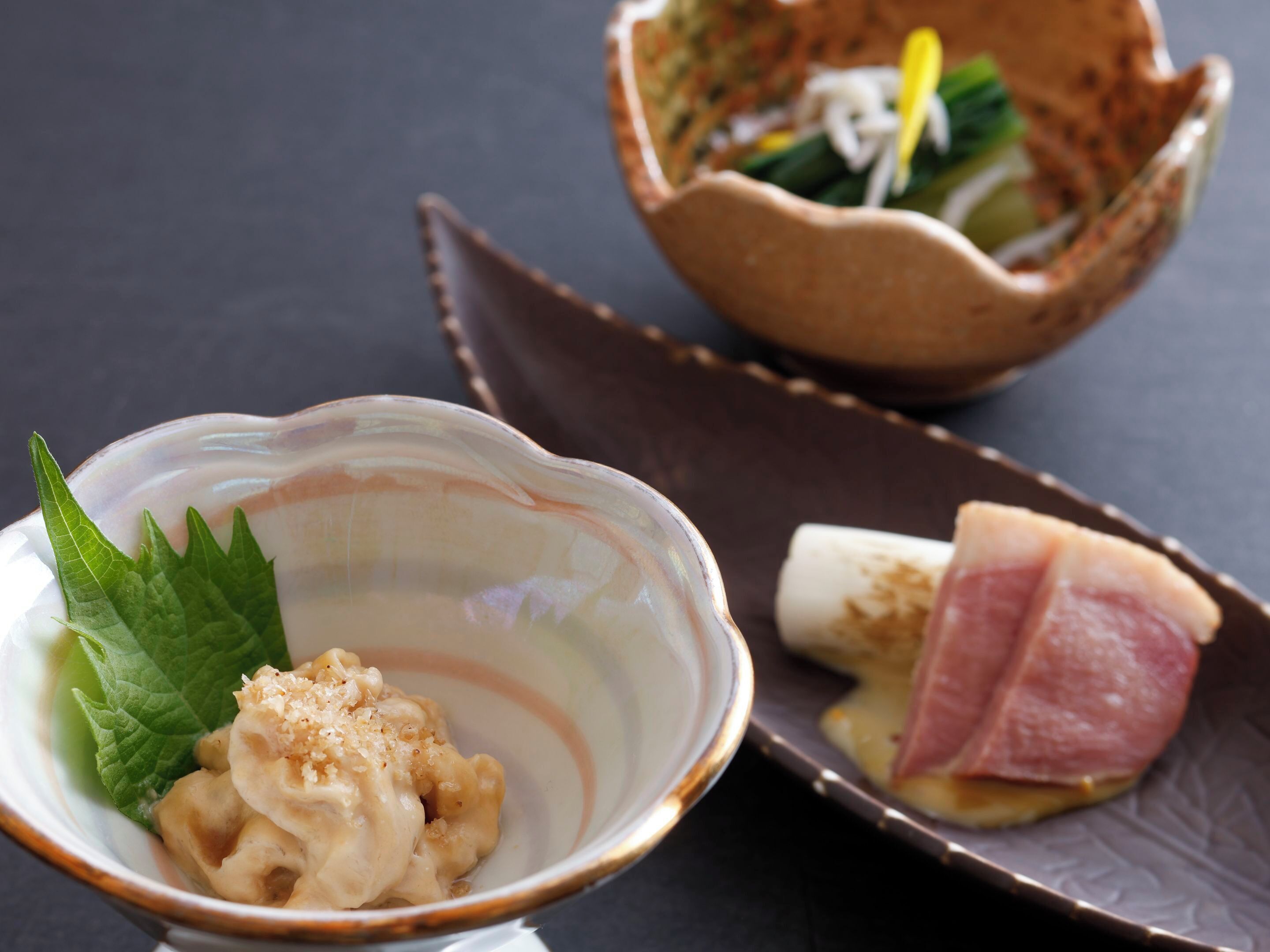 A bowl of food with a leaf garnish, a small dish with a leaf, and a plate with a slice of meat.