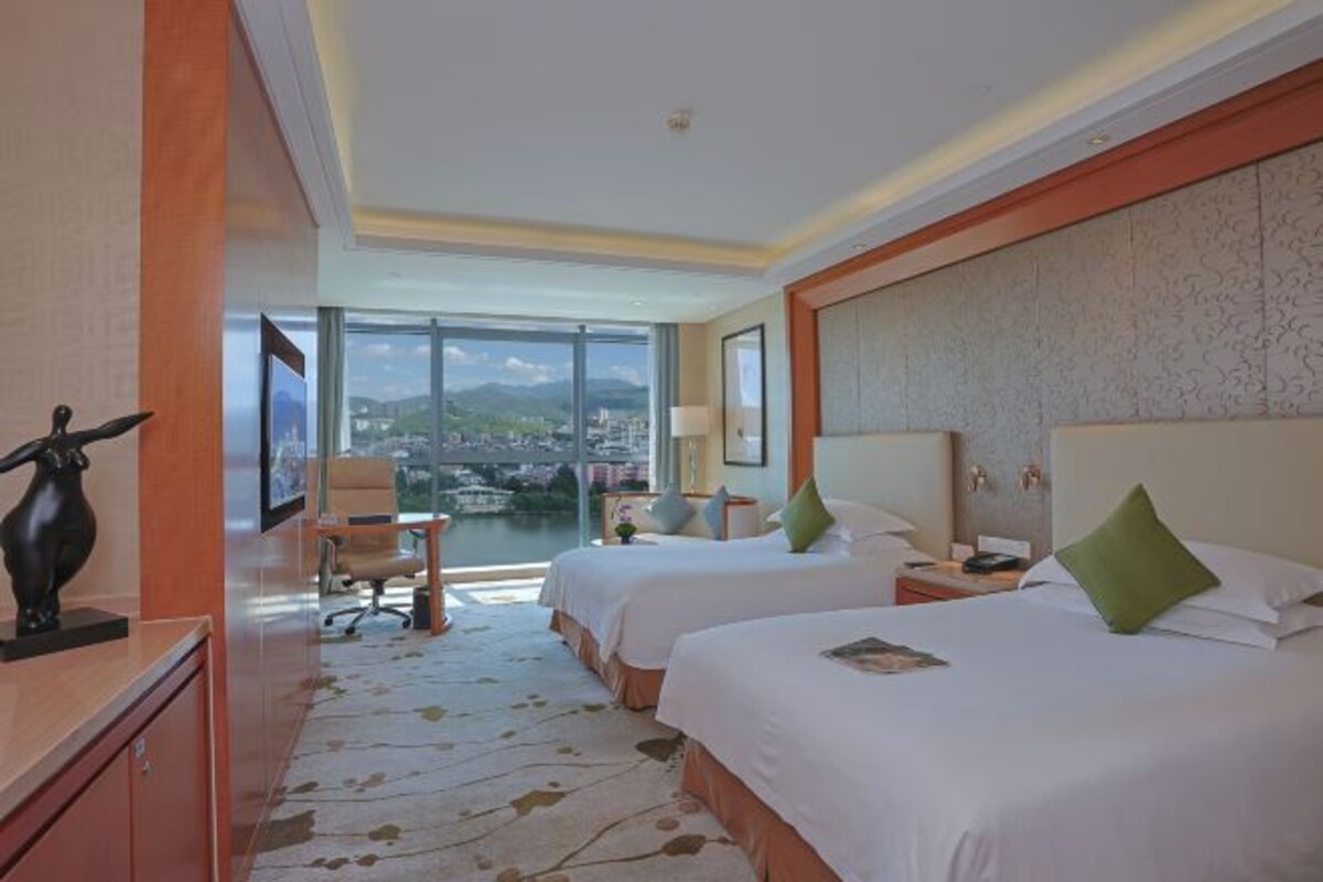 Quarto de hotel com duas camas, uma mesa, uma cadeira e vista para a paisagem urbana.