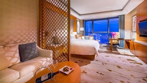 Une chambre d’hôtel avec un grand lit, un canapé, une table à manger avec un verre à vin, et une vue sur la ville à travers de grandes fenêtres.