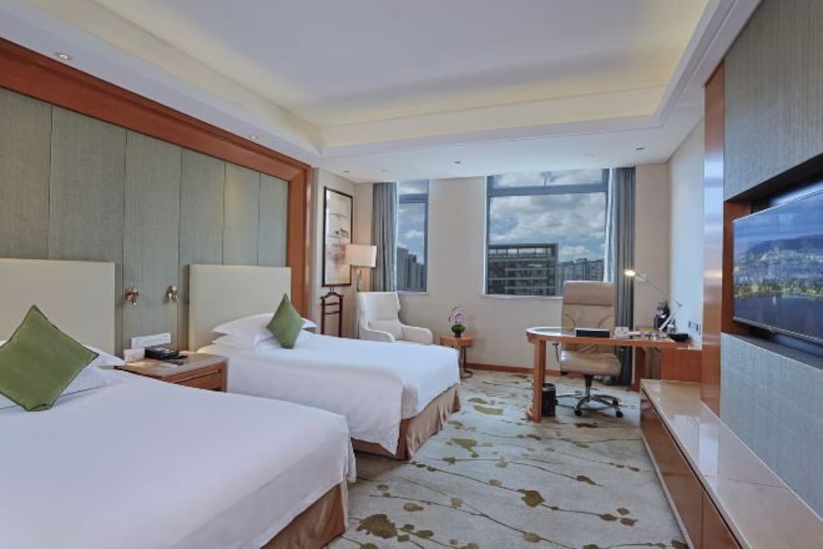Quarto de hotel com duas camas, uma escrivaninha, uma cadeira, televisão e uma janela grande com vista para a cidade.