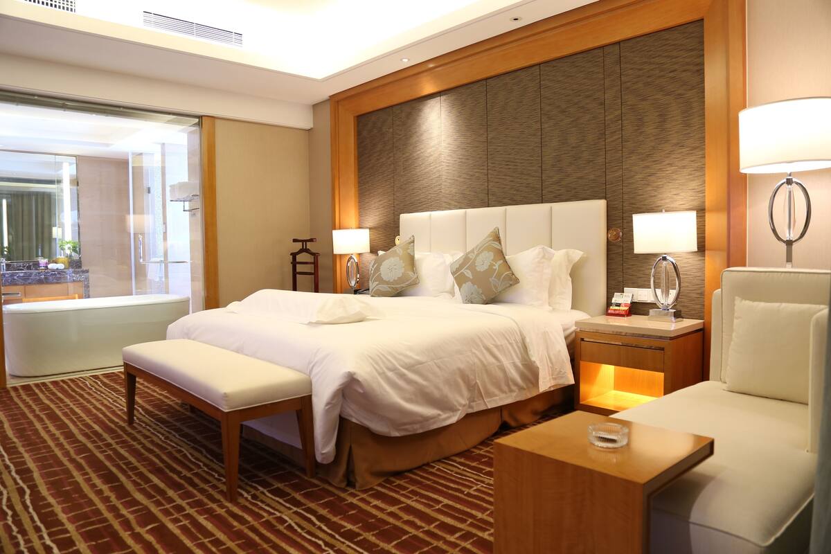Ein Hotelzimmer mit einem großen Bett, einem Nachttisch, einem Sessel und Blick ins Freie.