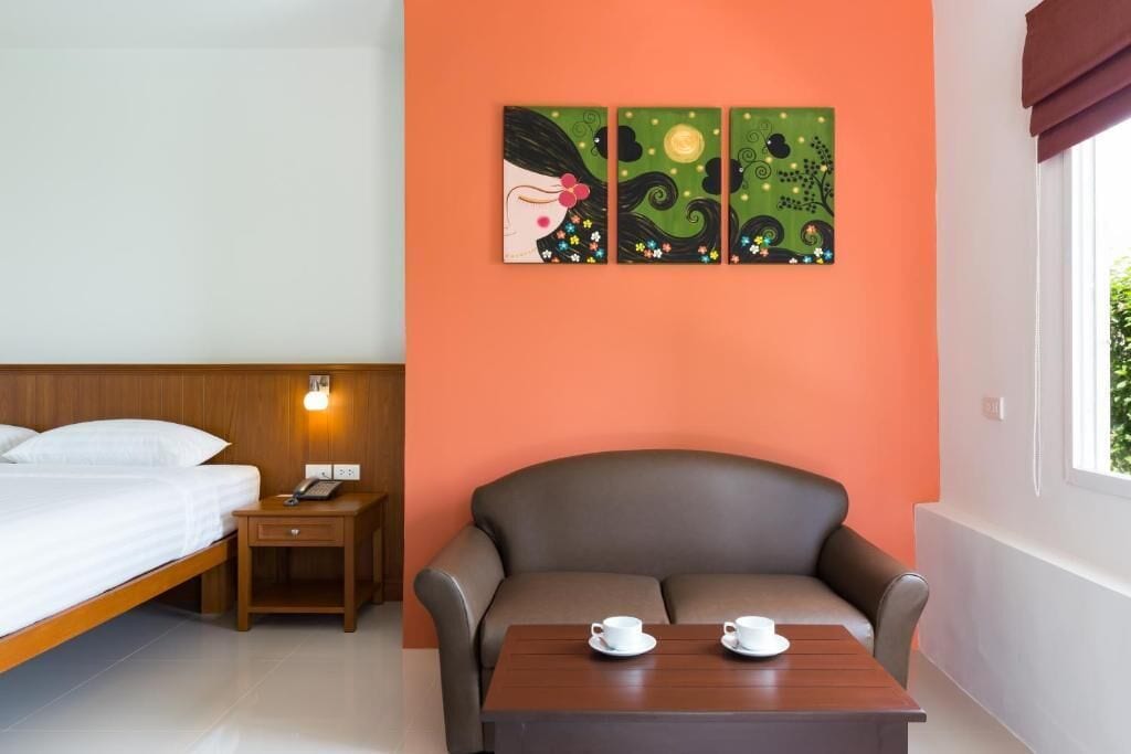 A hotel room with a bed, a nightstand with a lamp, a sofa, and a table with two cups.