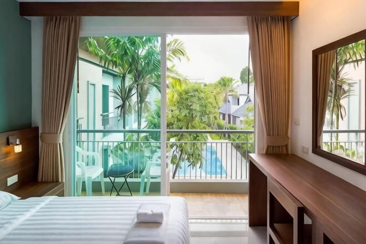 A hotel room with a bed, a desk, a chair, and a balcony with a view of greenery and a pool.