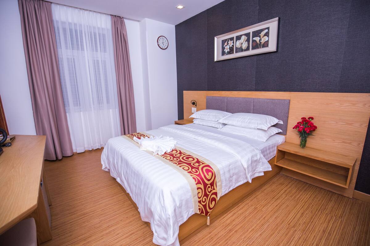 Ein ordentlich bezogenes Bett mit Holz-Kopfstück, ein Nachttisch mit Blumenvase und eine Uhr an der Wand.
