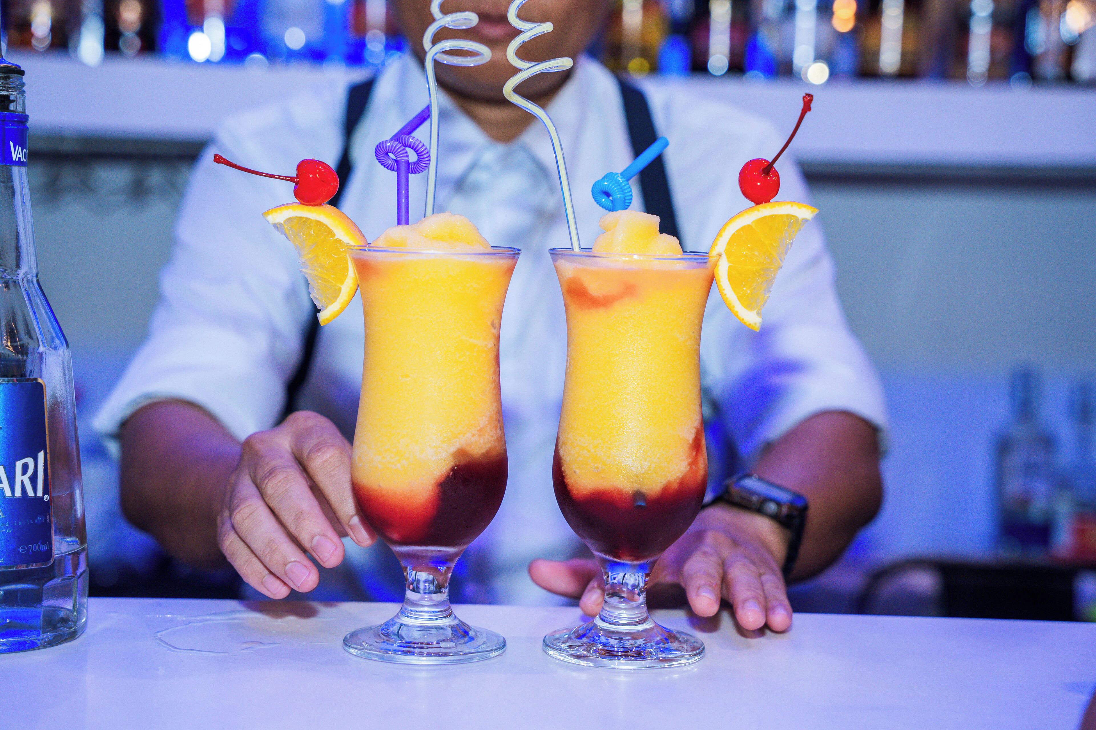 Two cocktails with fruit garnishes and straws, a bottle of water, and a bar setting.