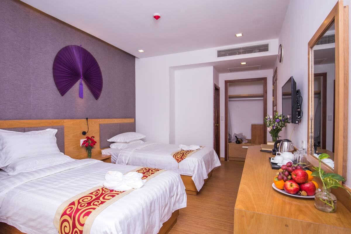 Ein Hotelzimmer mit zwei Betten, einem hölzernen Schreibtisch mit Obstschale, einem Spiegel und einem Wandventilator.