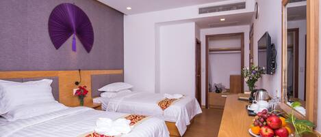 A hotel room with two beds, a wooden desk with a fruit bowl, a mirror, and a wall fan.