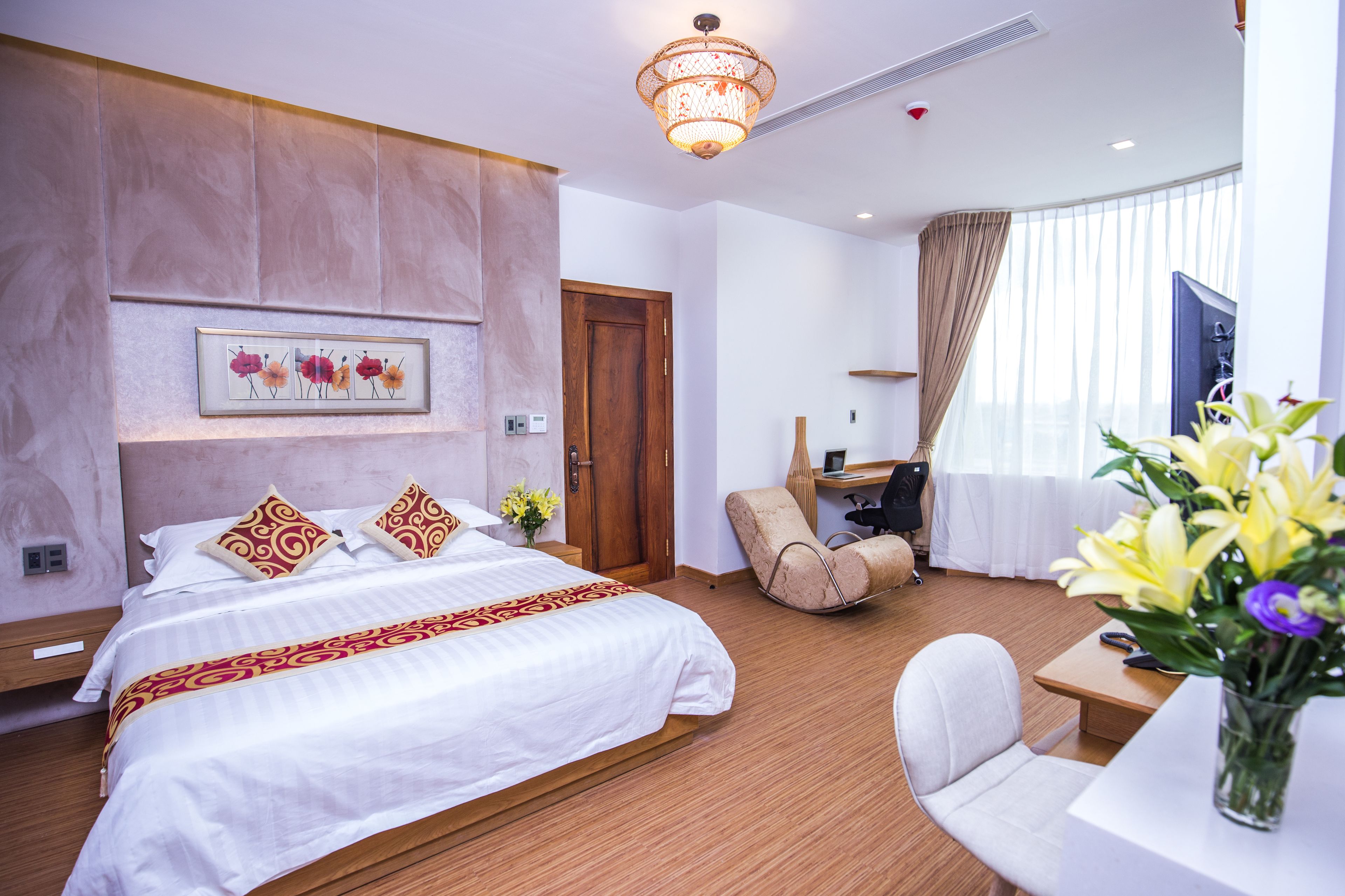 Habitación de hotel con una cama grande, un escritorio, una silla, una lámpara de escritorio, un florero con flores y una ventana con cortinas.