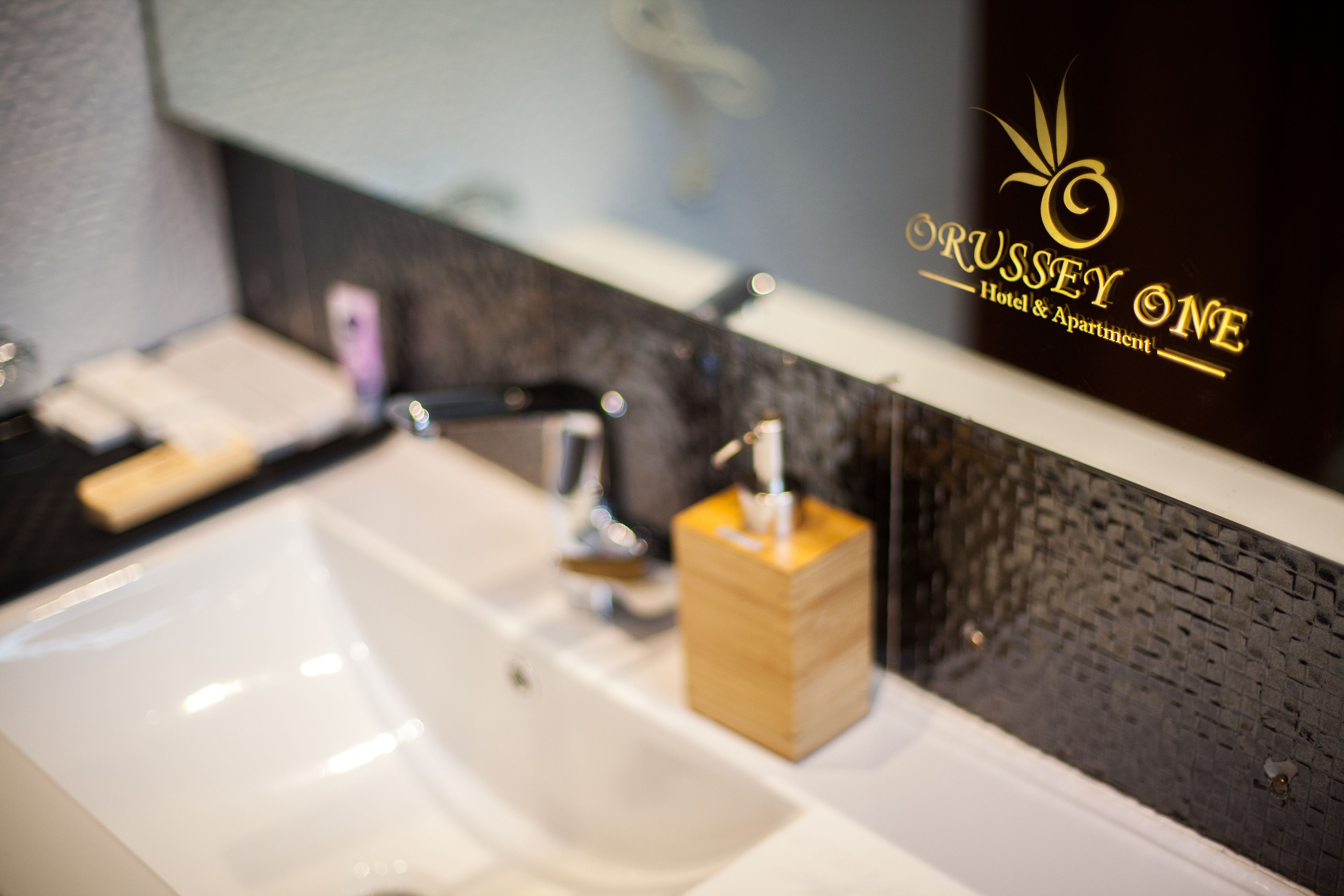 Un bagno con lavandino, specchio e dispenser per sapone in legno. Lo specchio riflette un cartello con su scritto "Russsey One Hotel & Apartment".