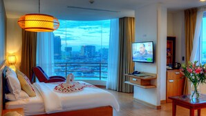 Habitación de hotel con una cama grande, televisor, escritorio con un jarrón de flores y vistas a la ciudad a través de amplios ventanales.