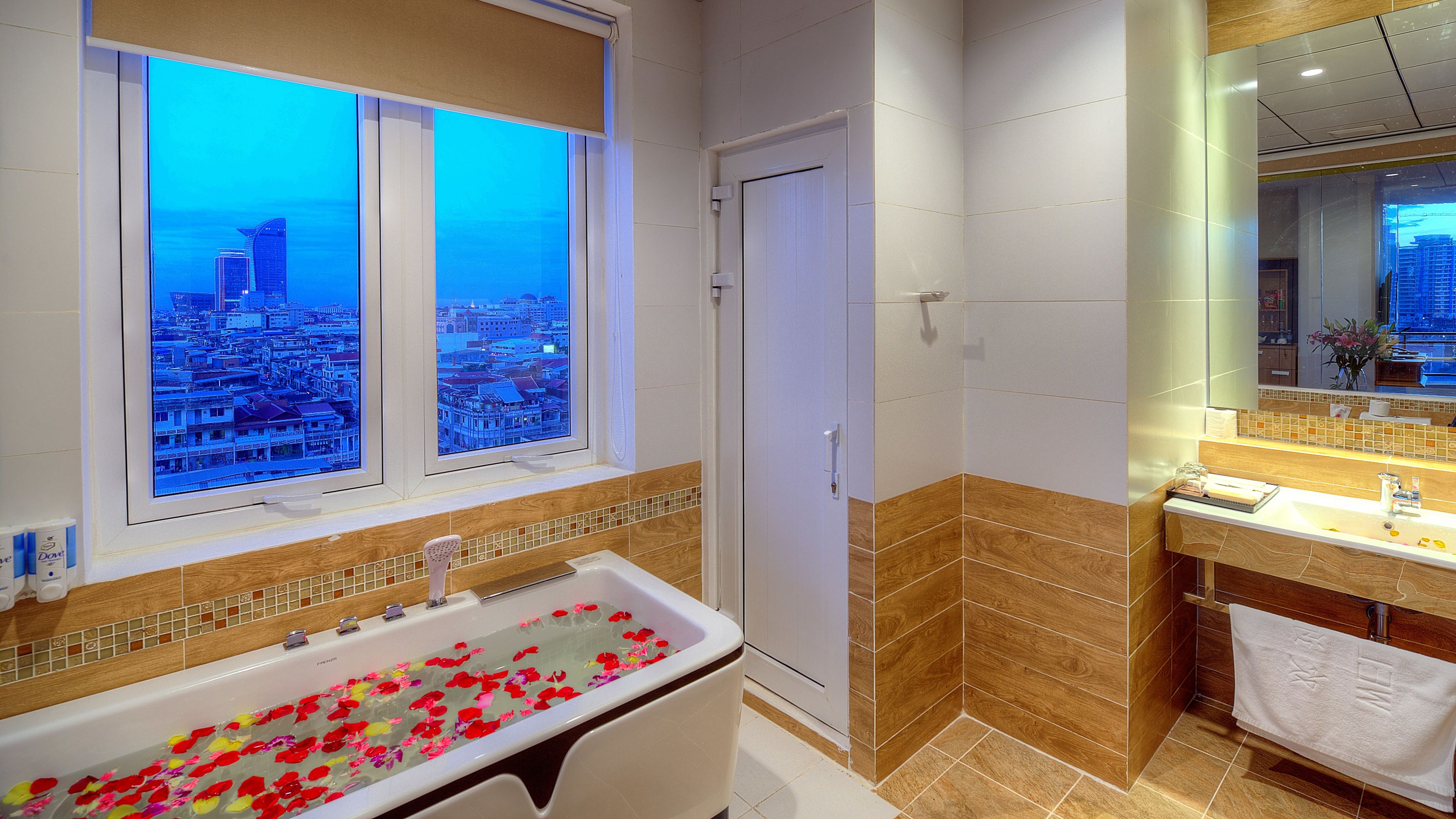 A modern bathroom with a bathtub filled with rose petals, a large window with a city view, and a wooden vanity with a mirror.
