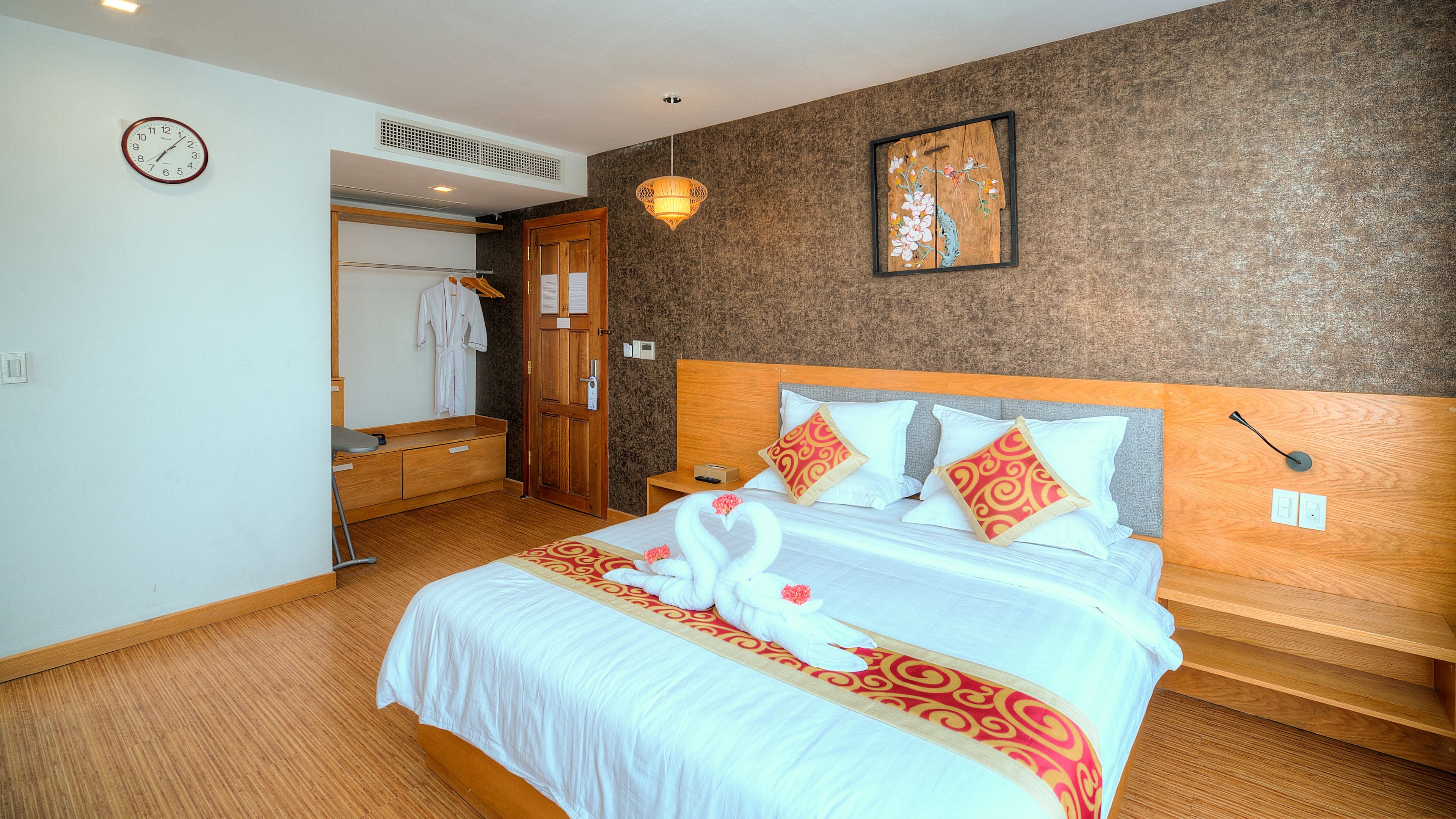 A hotel room with a bed, a wooden headboard, a nightstand, a wall clock, a robe rack, and a painting.