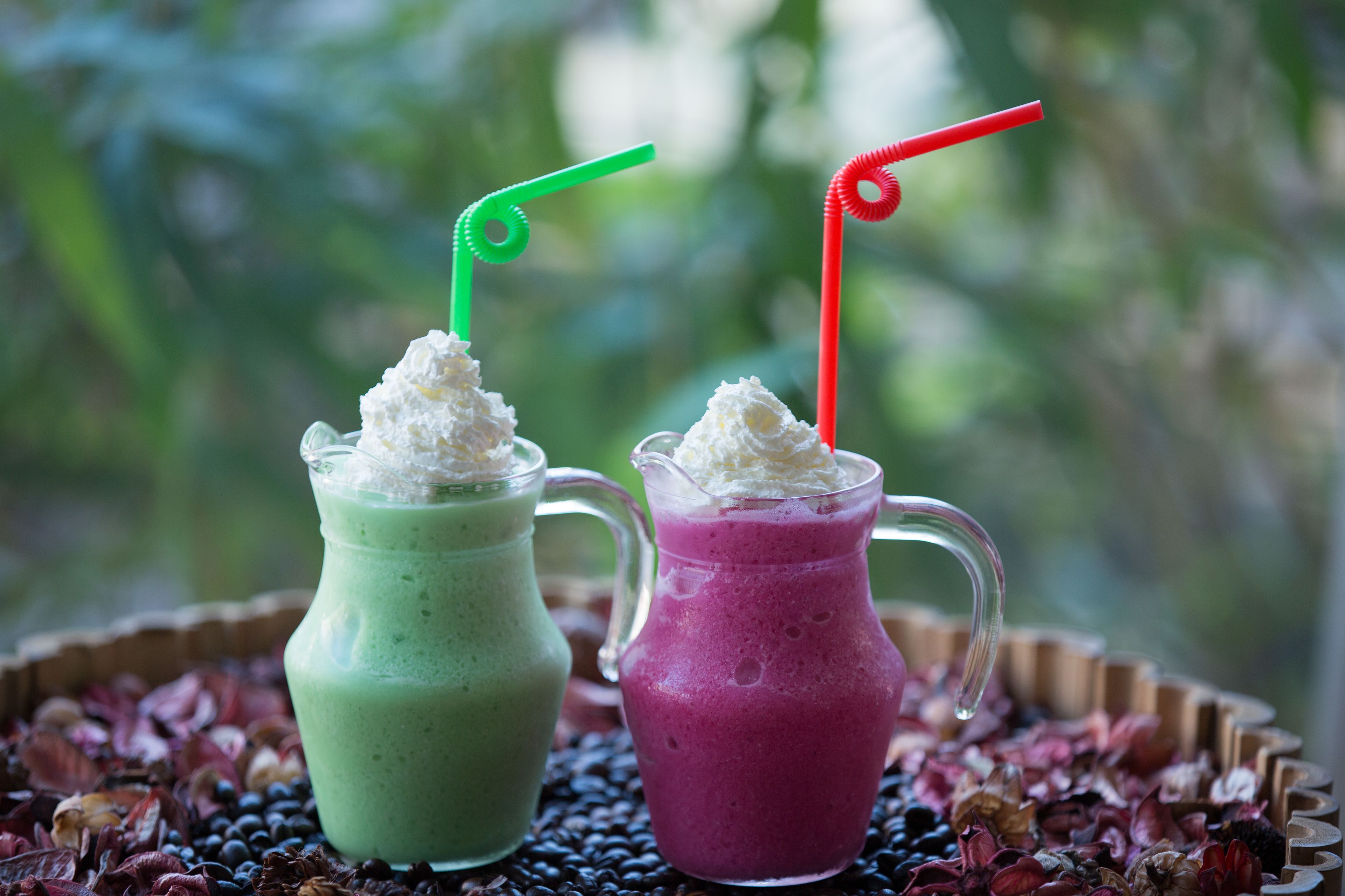 Two colorful smoothies with whipped cream and straws, served in glasses, on a bed of dried fruit.
