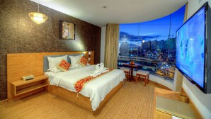 Habitación de hotel con cama, mesita de noche, una pequeña mesa con una silla, un ventanal con vistas a la ciudad y un televisor de pantalla plana.