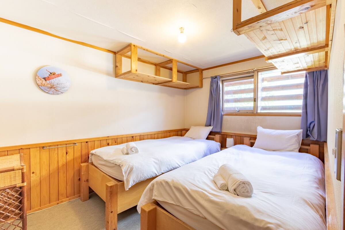 A room with two wooden beds, a wooden bench, a small table, and a wall-mounted shelf.