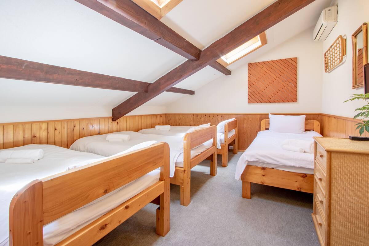A room with four wooden beds arranged in a corner, a small wooden dresser, and a sloped ceiling with wooden beams.