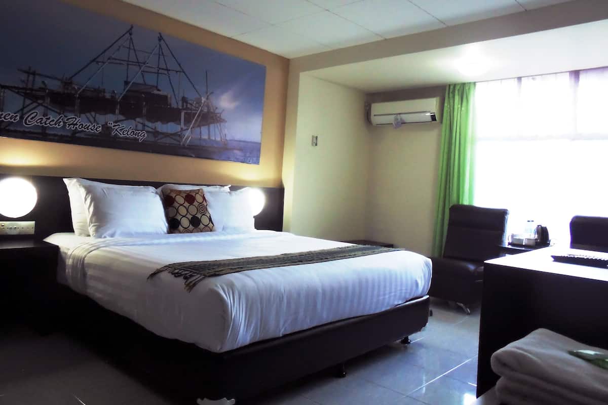 A hotel room with a large bed, a desk, and a chair. There is a painting on the wall and an air conditioning unit on the wall.