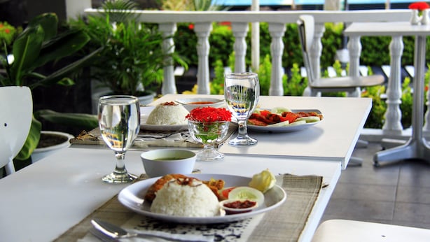 Mesa posta com arroz, vegetais e um copo de água, com vista para o verde e um corrimão ao fundo.
