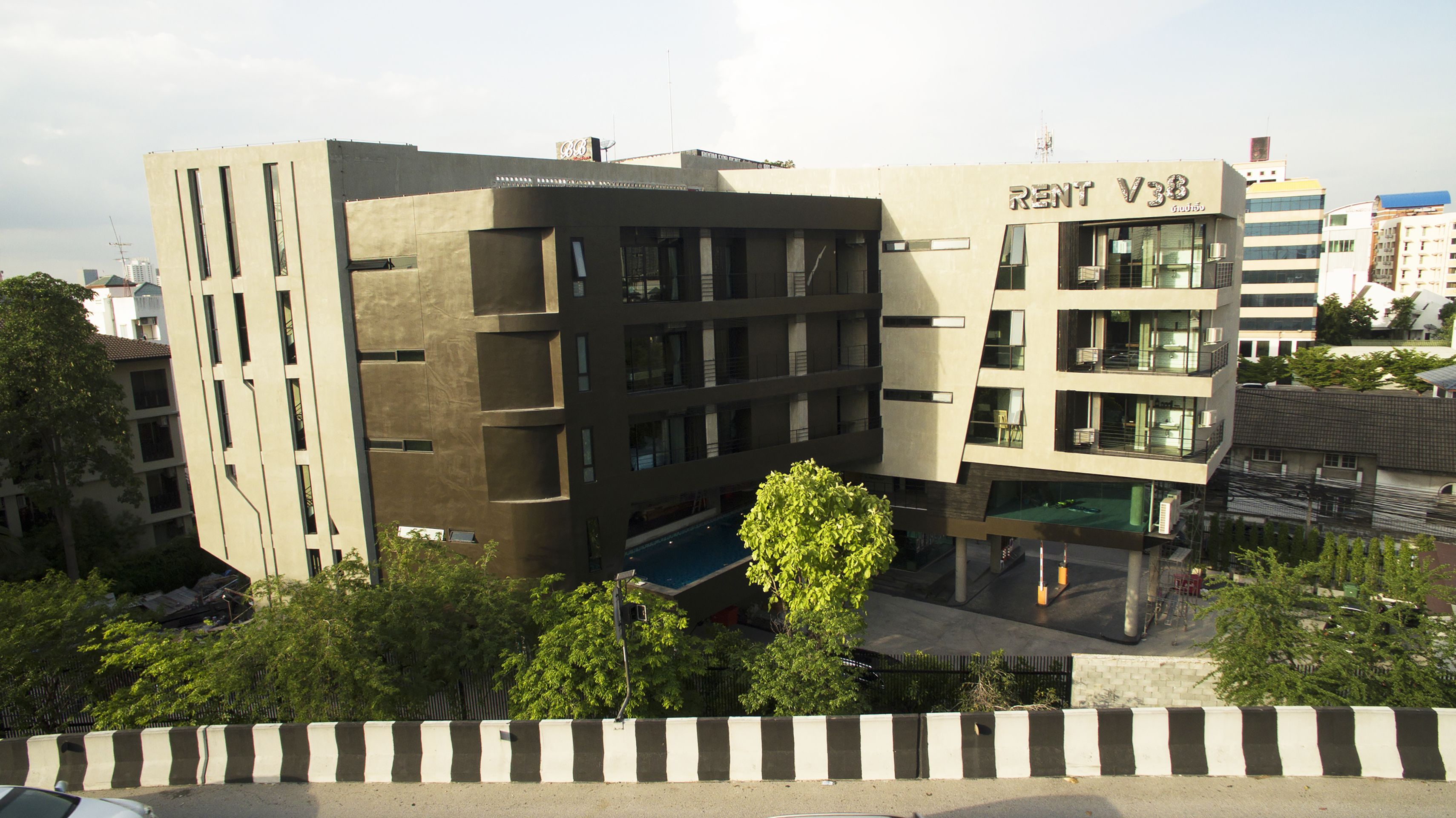 A modern building with a 'RENT V3' sign, featuring a flat roof, multiple windows, and a visible pool area.