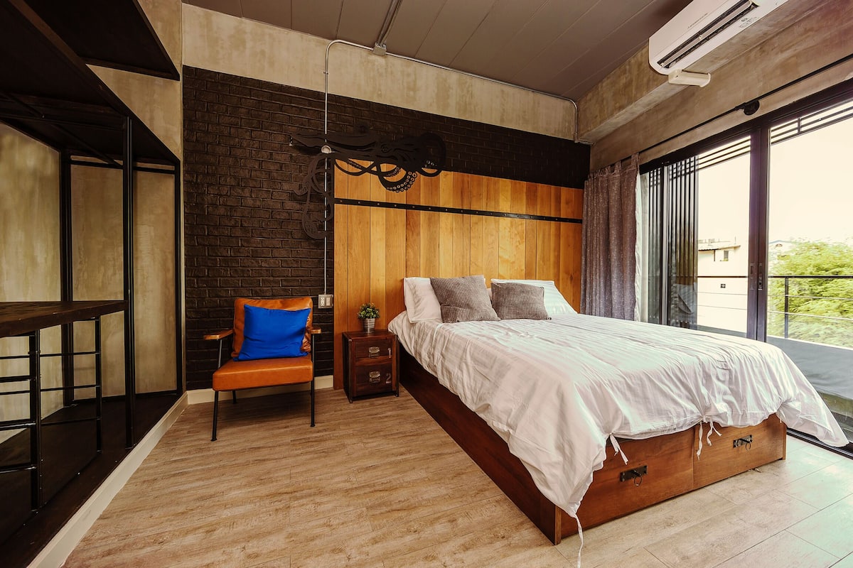 A modern bedroom with a large bed, a wooden nightstand, a chair, and a view of the outdoors through a large window.