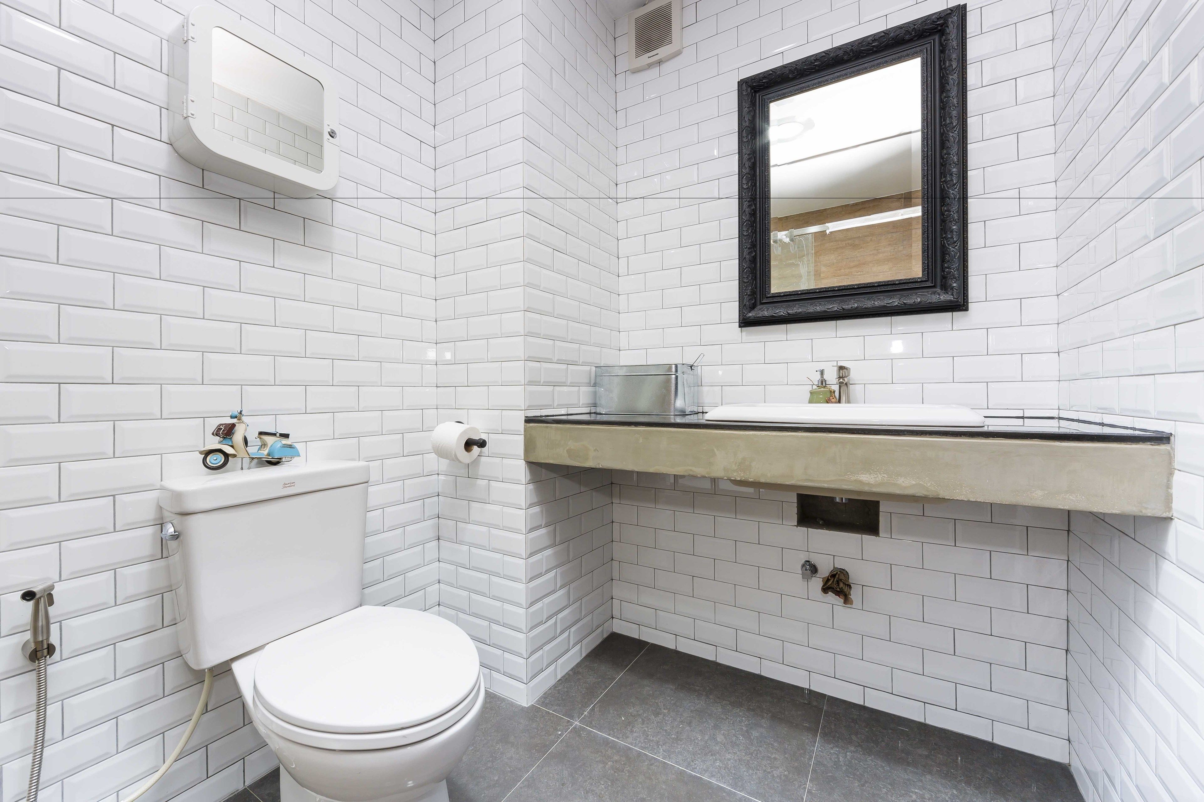 Une salle de bain avec des carreaux de métro blancs, des toilettes blanches, un lavabo avec un plan de toilette beige et un miroir à cadre noir.