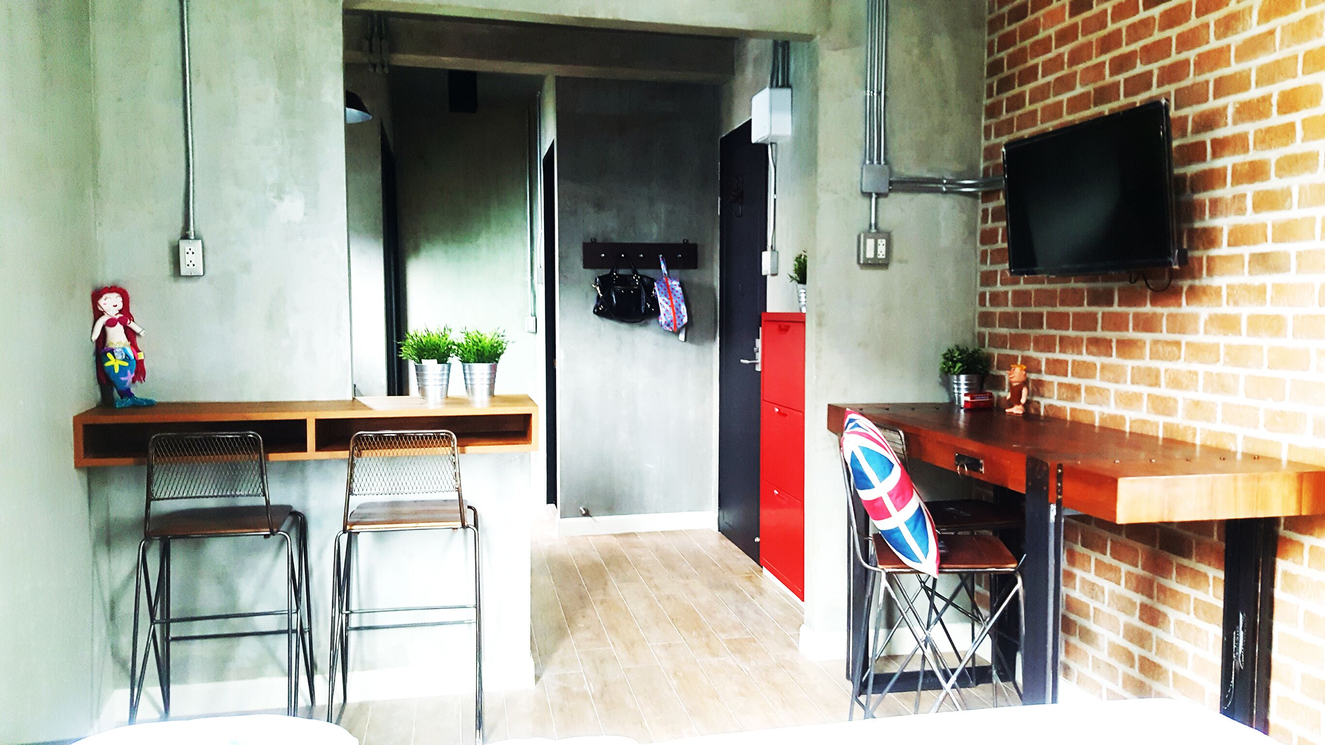 A compact kitchen with a wooden countertop, two metal chairs, a red cabinet, and a TV mounted on a brick wall.