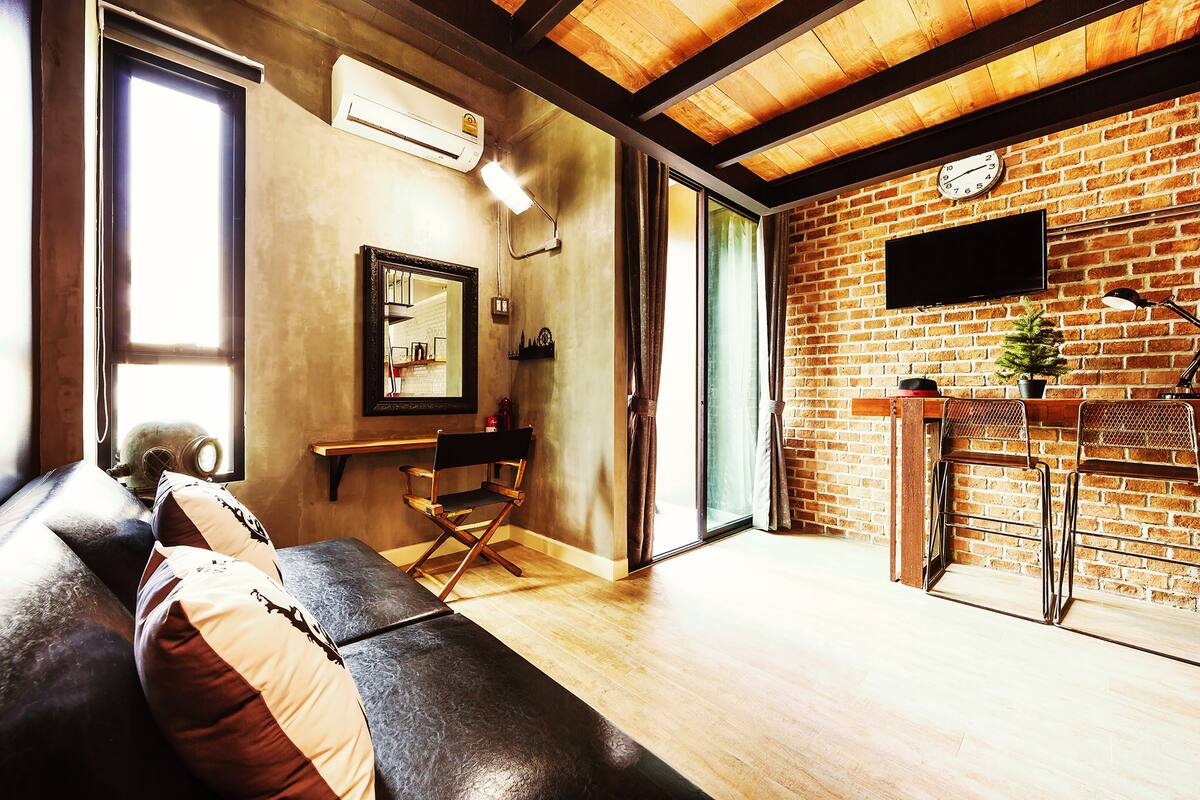 A room with a brick wall, a flat-screen TV, a wooden ceiling, a leather sofa, and a sliding glass door.