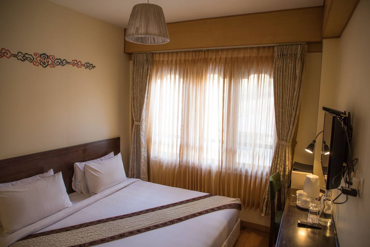 A hotel room with a bed, a desk with a lamp, a television, and a window with curtains.