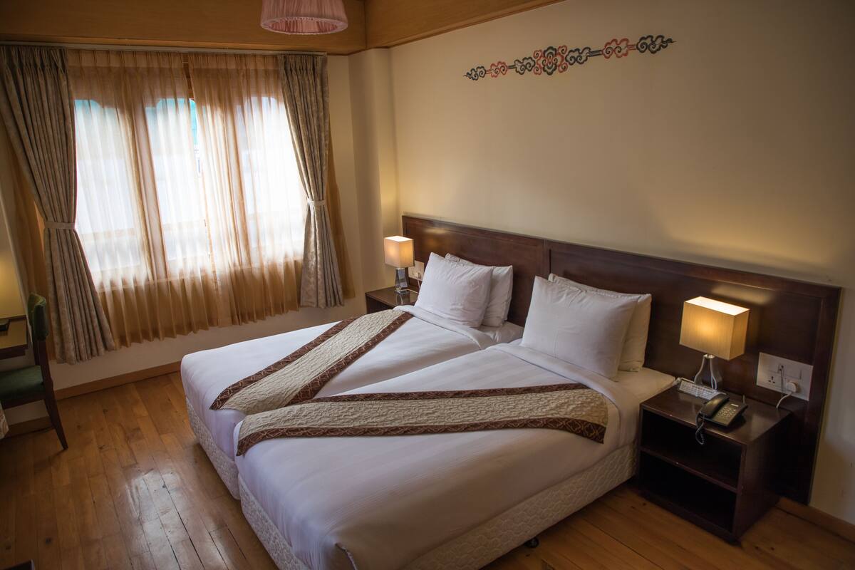 A double bed with white linens and brown decorative borders, two bedside lamps, a wooden nightstand with a phone, and a window with sheer curtains.