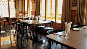 Een restaurant met meerdere tafels en stoelen, grote ramen voorzien van gordijnen, en een gepolijste vloer.
