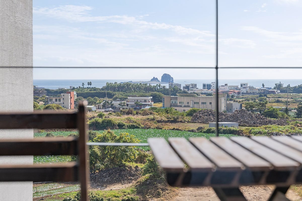 A view from a balcony with a wooden bench, overlooking a coastal town with buildings and a rocky outcrop in the distance.