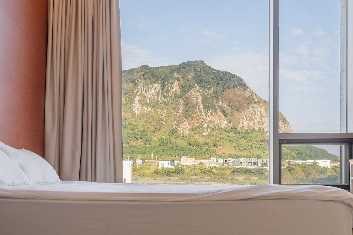 A hotel room with a bed, a window with curtains, and a view of a mountain.