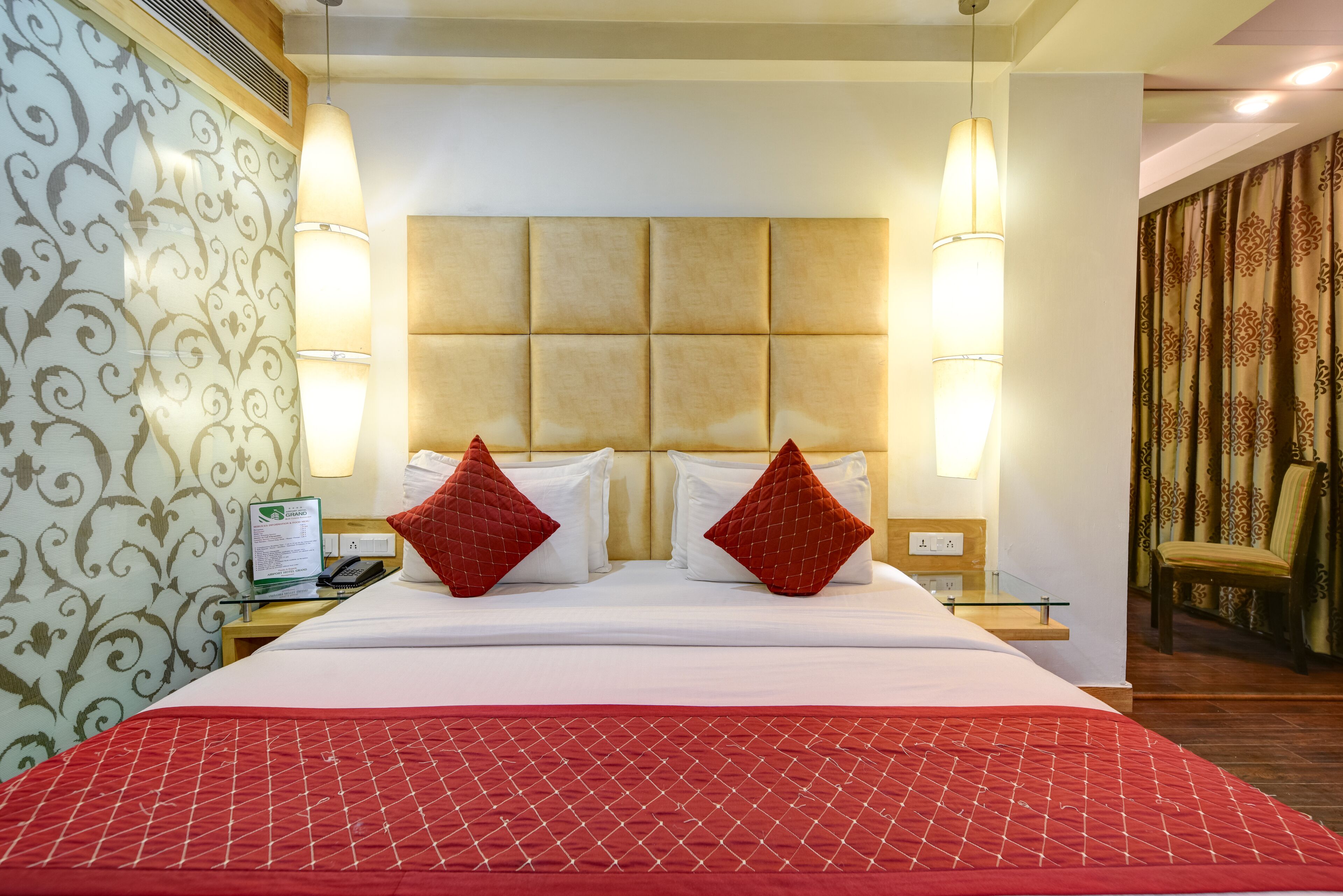 A hotel room with a large bed, two red pillows, a wooden headboard, bedside lamps, a nightstand, and a chair.
