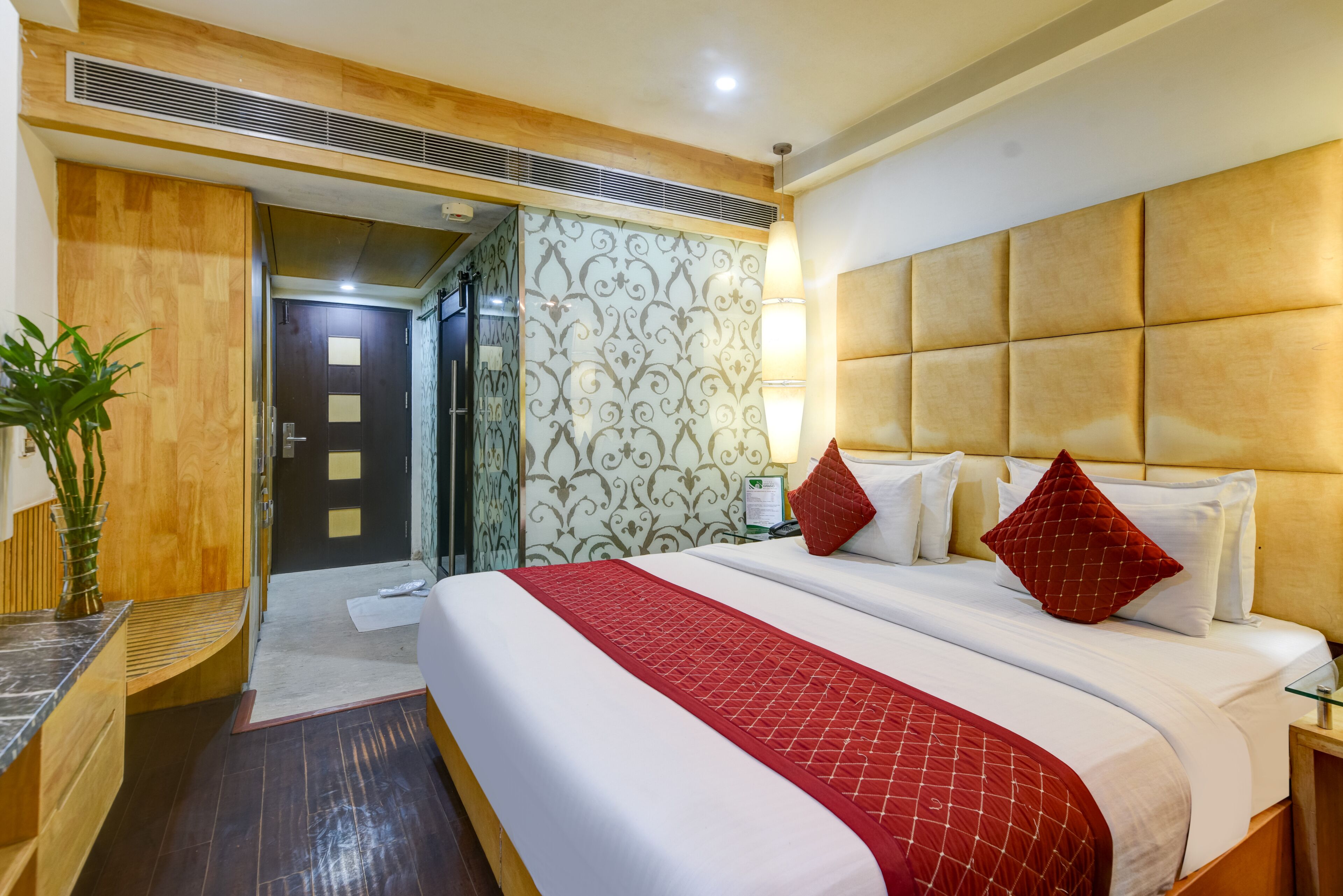 A hotel room with a large bed, a wooden headboard, a red and white bedspread, and a potted plant.