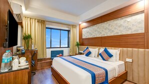 Una habitación de hotel con una cama grande, cabecera de madera, un escritorio con televisor de pantalla plana, una ventana con cortinas y una planta.