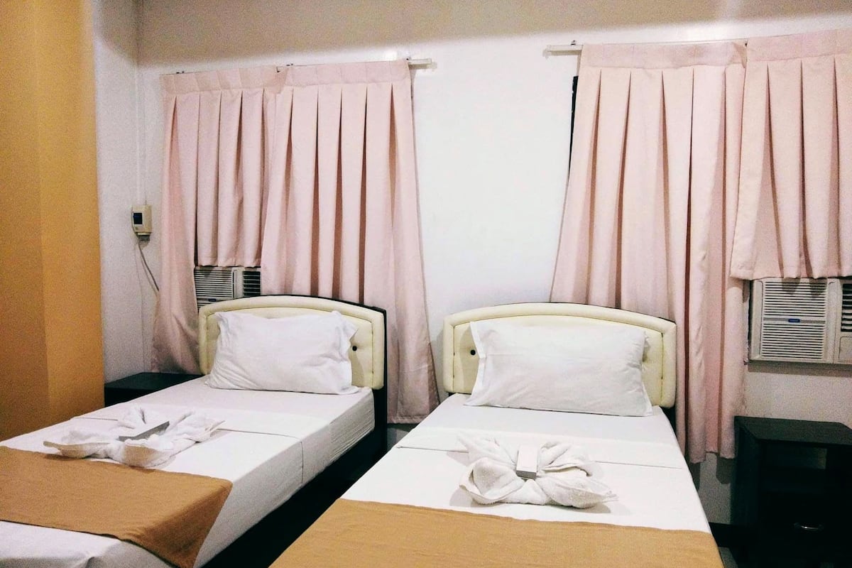 A hotel room with two single beds, each with a white pillow and a brown blanket. There are pink curtains on the windows, and an air conditioning unit is visible on the wall.