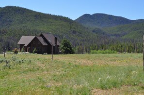 A rustic cabin with a sloped roof, surrounded by a grassy field and dense forest. - Homestake Lodge (Whitehall)