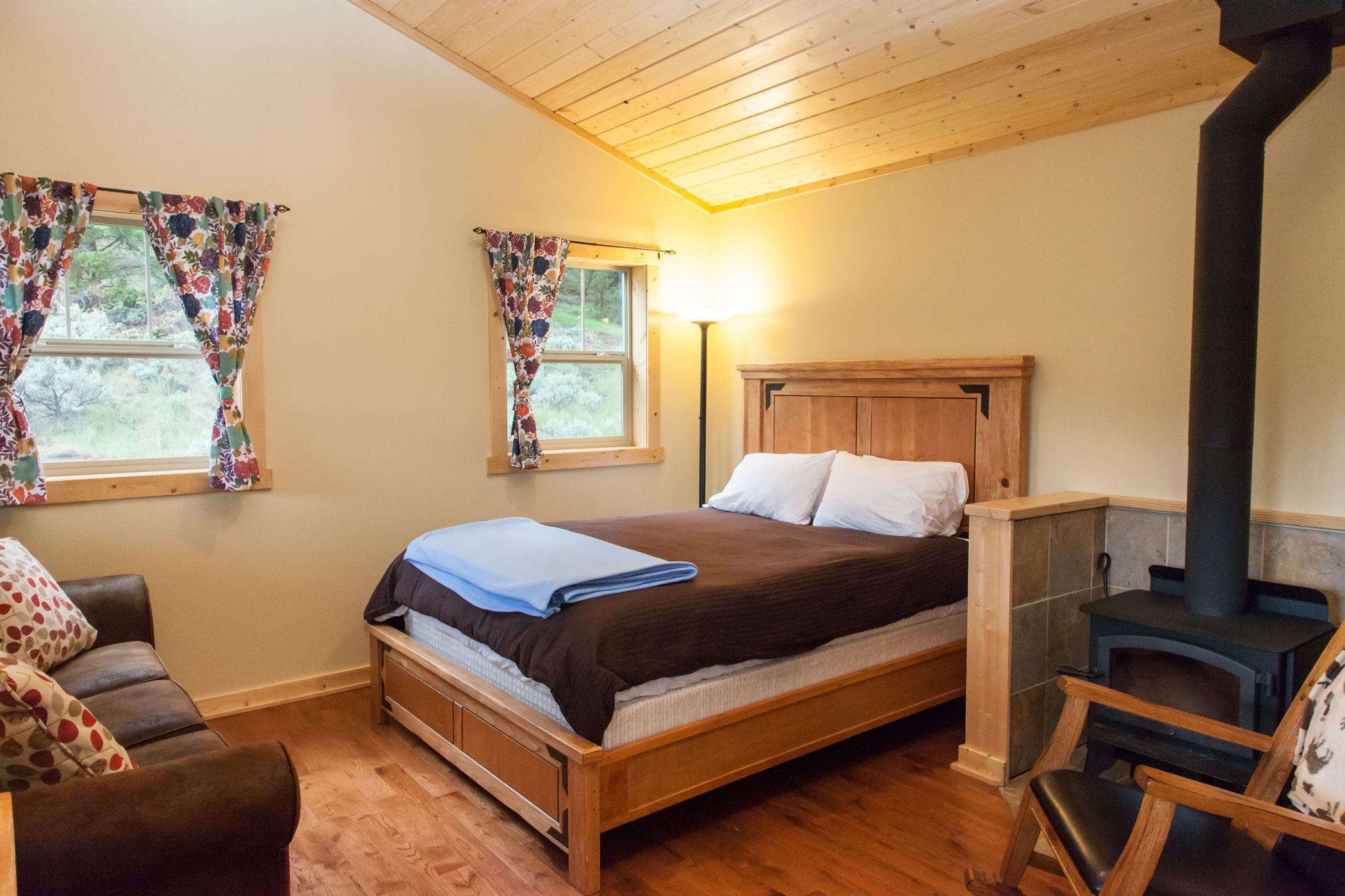 Une chambre confortable avec un lit en bois, deux fenêtres ornées de rideaux à motifs floraux, une chaise en bois et un poêle à bois.
