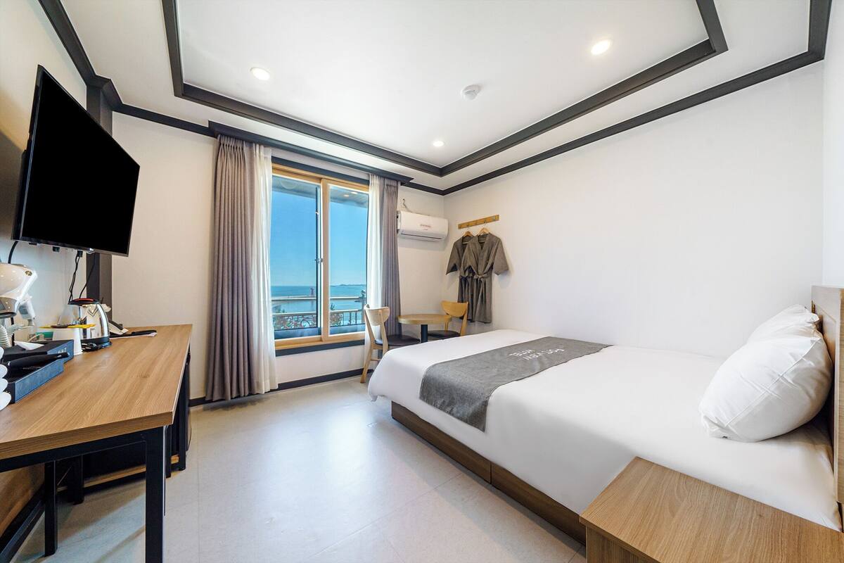 A hotel room with a bed, a desk, a TV, and a view of the sea.