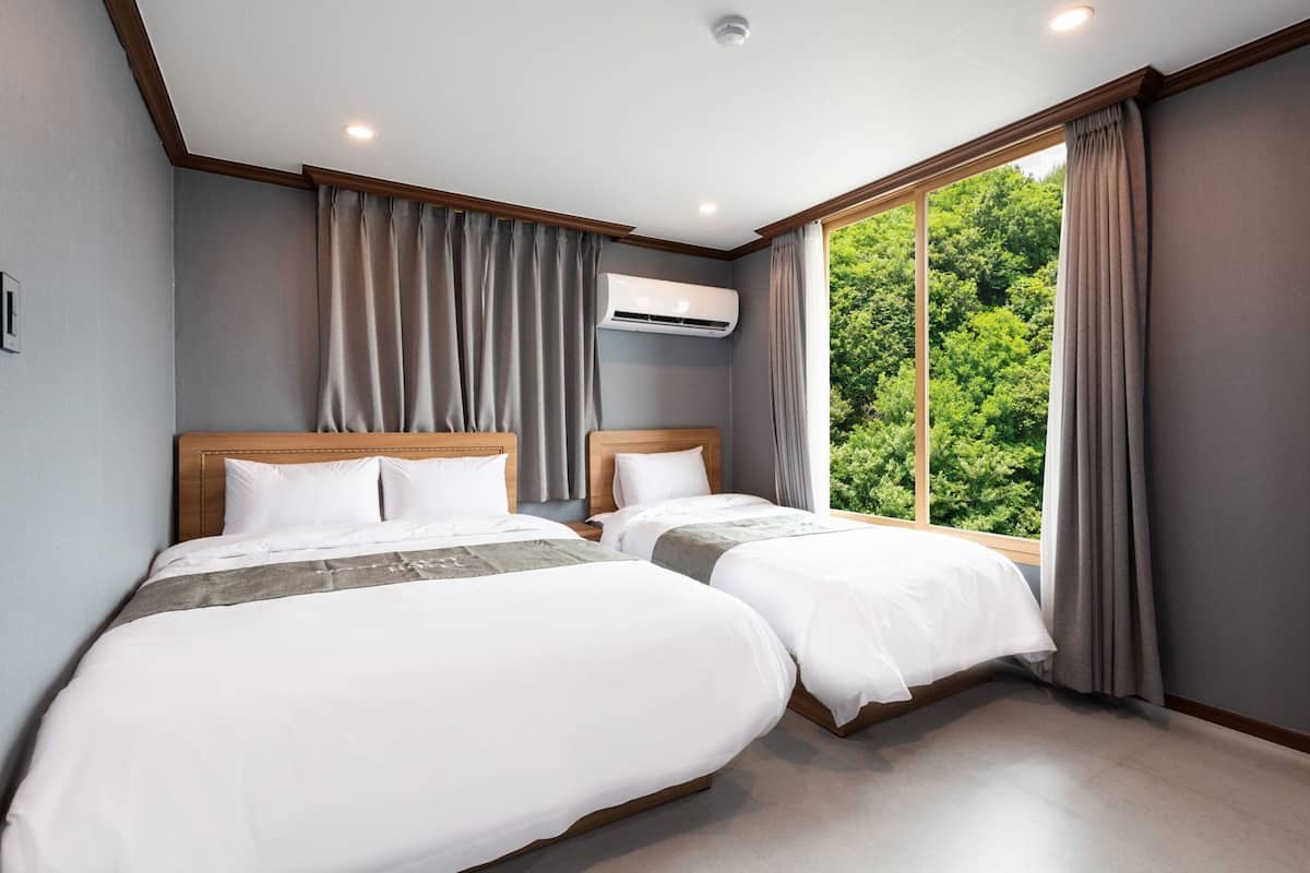 A hotel room with two beds, a large window with curtains, an air conditioning unit, and a ceiling light.