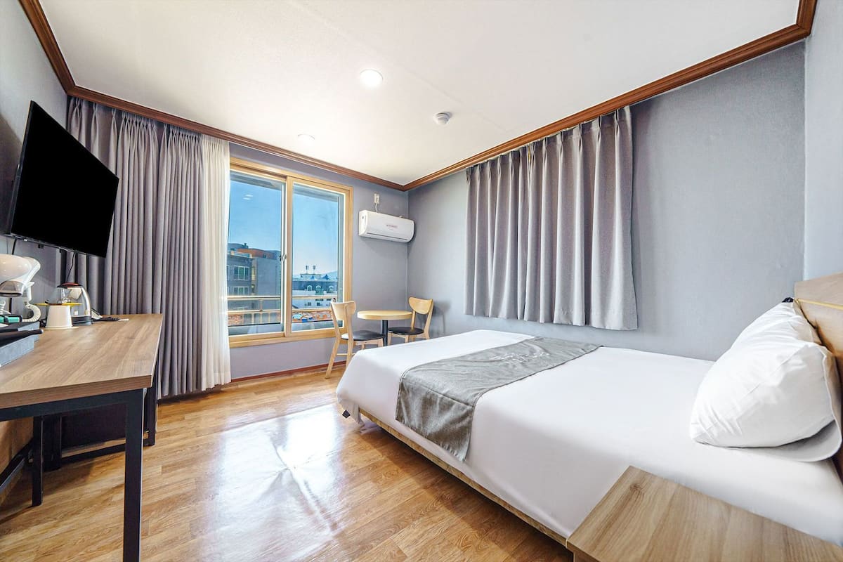 A hotel room with a bed, a desk with a computer, a chair, a TV, and a window with curtains.