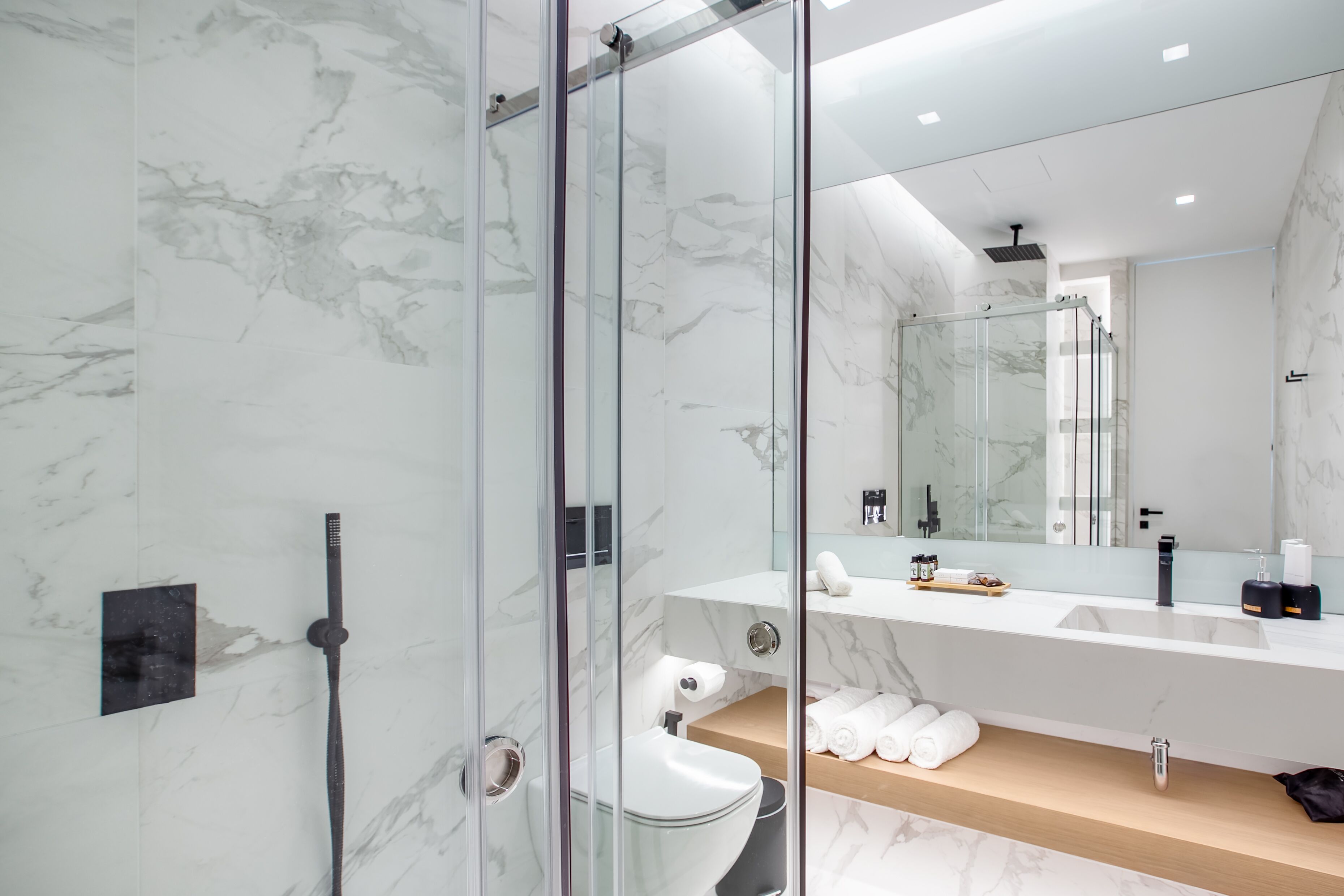 A modern bathroom with a glass-enclosed shower, a marble wall, a large mirror, and a wooden vanity with towels.