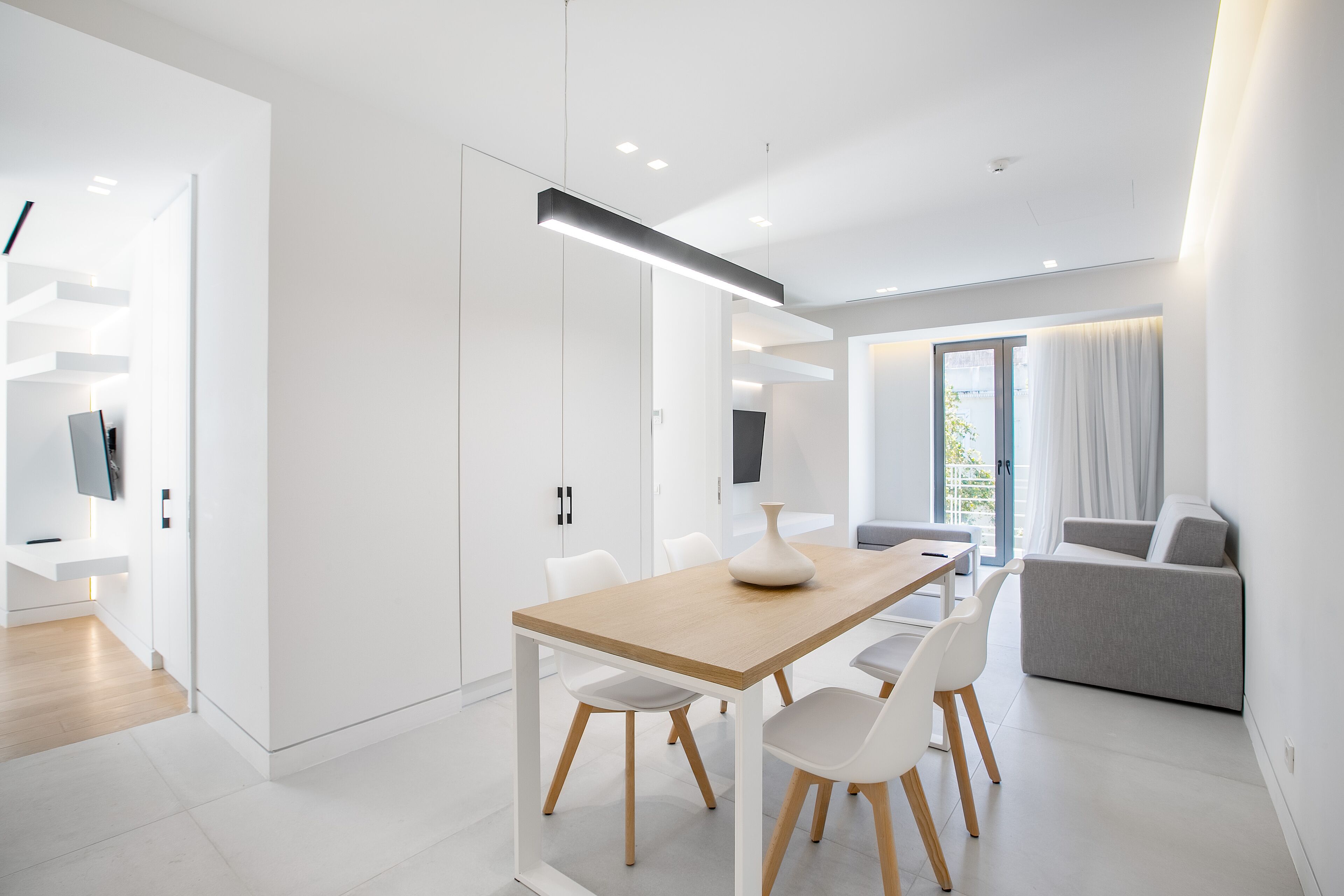 A modern, minimalist dining area with a wooden table and white chairs, a grey sofa, and a flat-screen TV mounted on the wall.