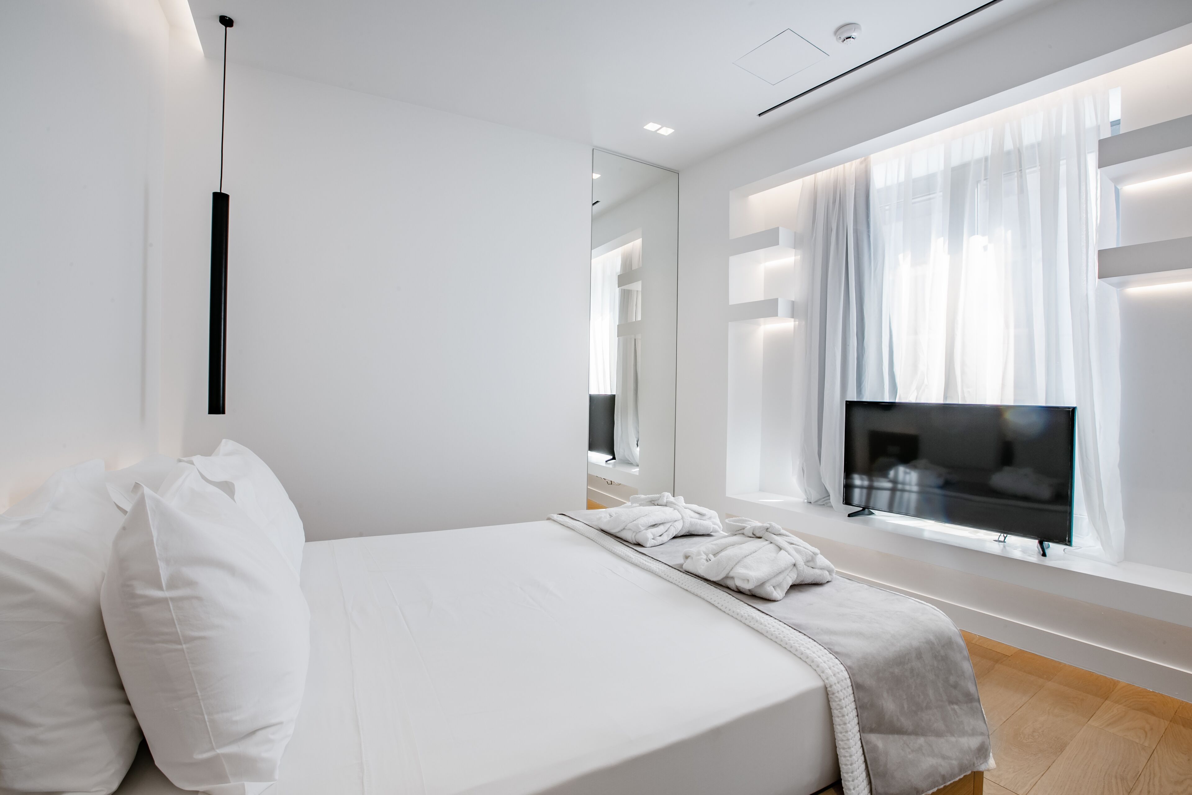 A modern hotel room with a large bed, a flat-screen TV, a large window with sheer curtains, and a mirror on the wall.